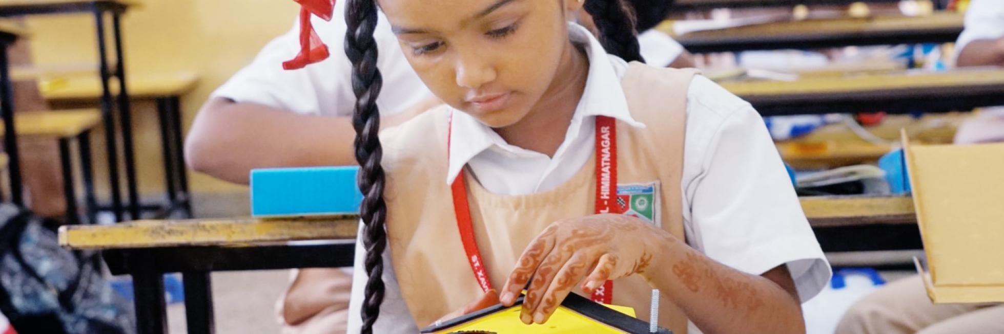 A young girl in school uniform focuses intently while working on a yellow model house in a classroom setting.