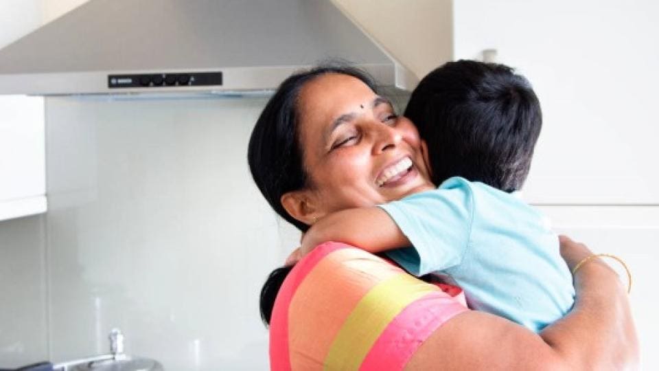 A woman happily embraces a child in a modern kitchen, with a stovetop and range hood in the background.