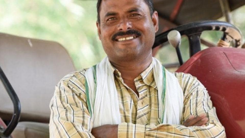 A man in a striped shirt and white scarf stands smiling with arms crossed in front of a red tractor.